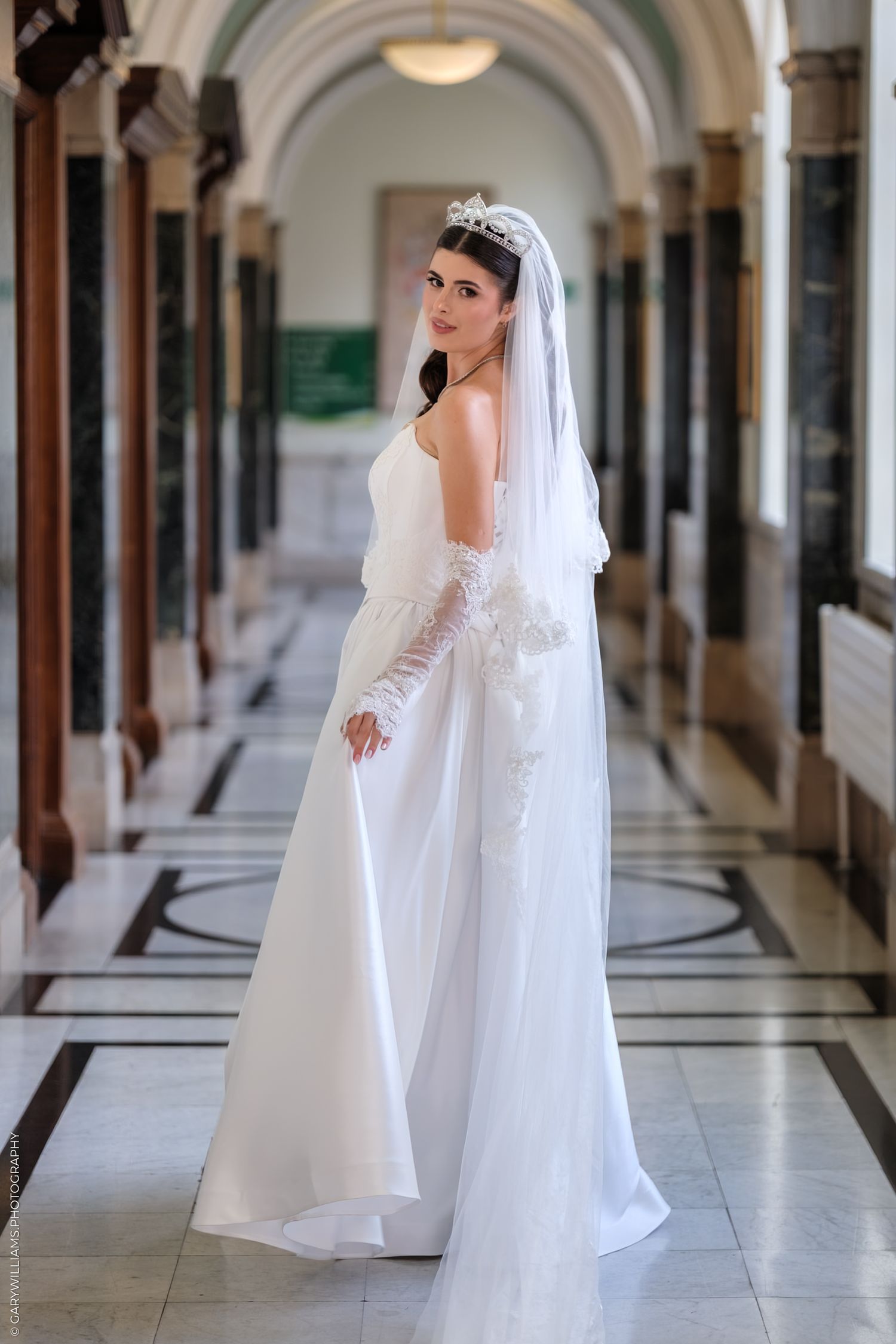A bride in a white wedding gown and veil poses in an elegant hallway with repeated archways and columns.