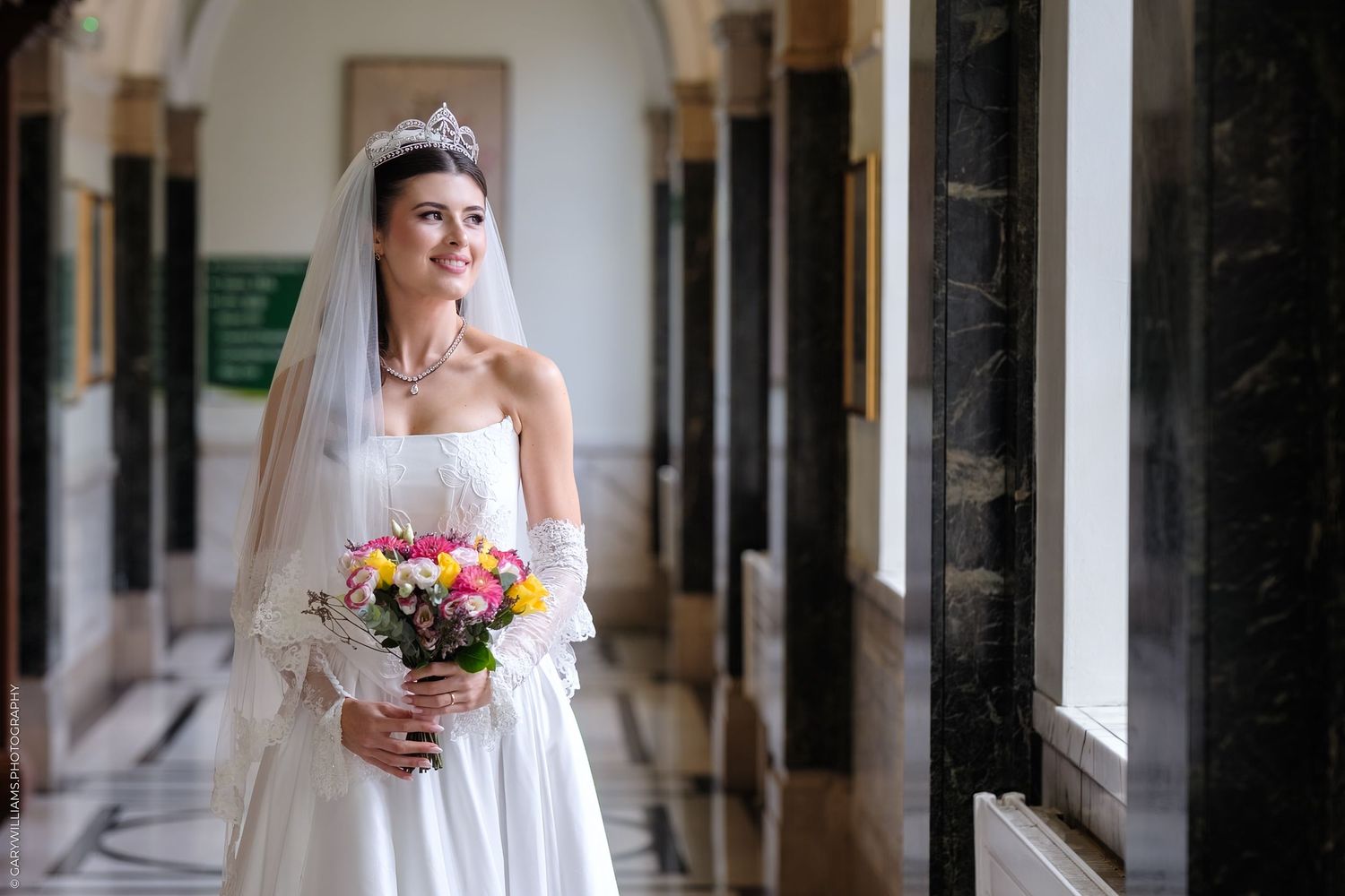A bride in a traditional white wedding dress and veil holds a vibrant bouquet while standing in an elegant columned hallway.