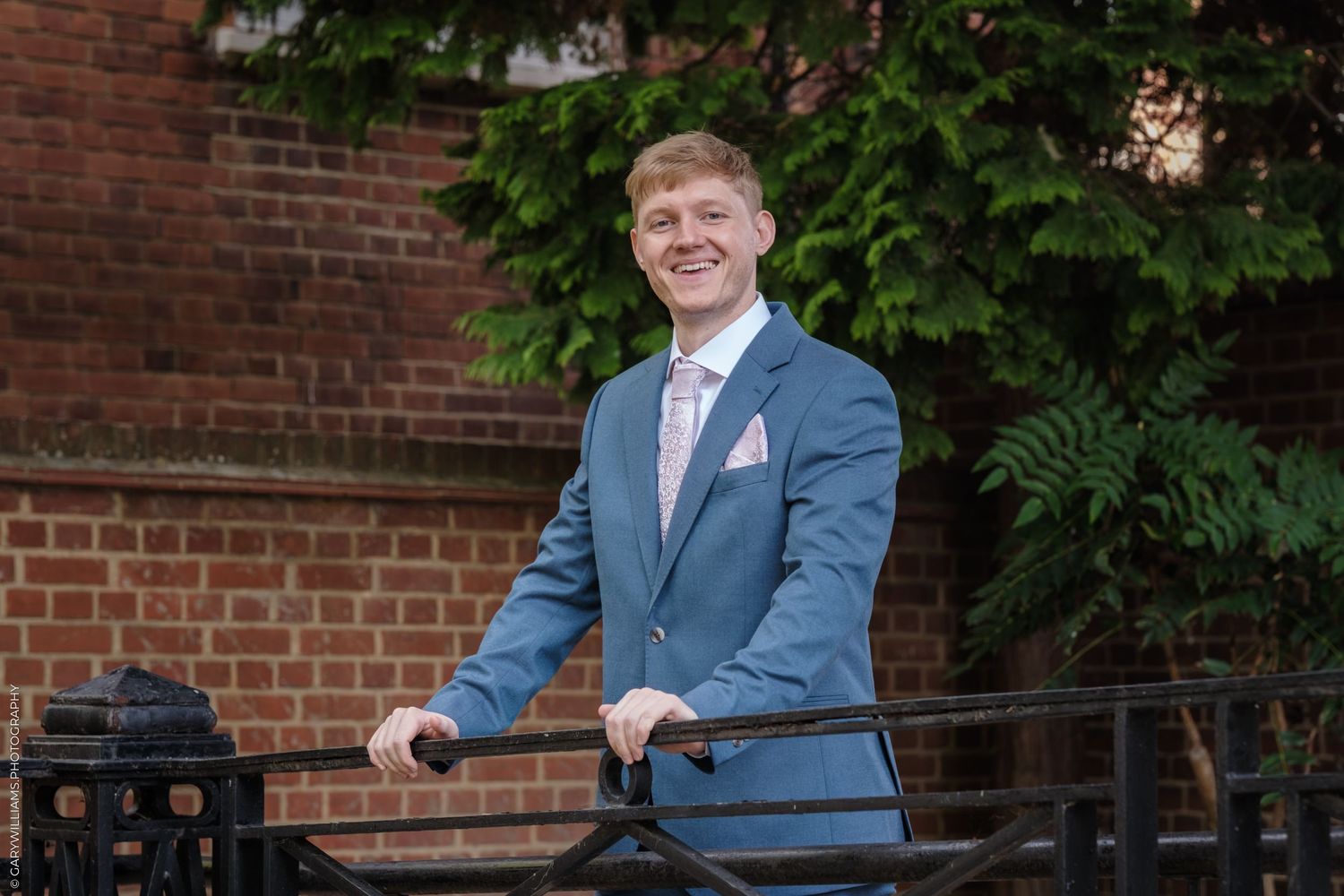 A person wearing a light blue suit and pink tie poses against a brick wall with greenery.