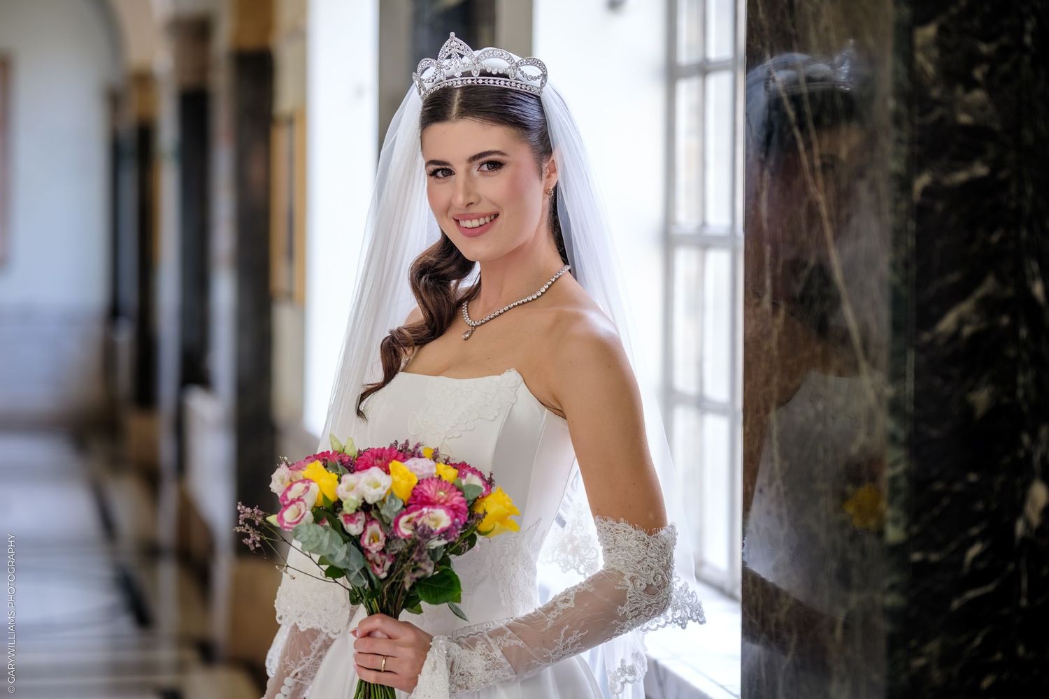 Bride in strapless white wedding gown holds colorful spring bouquet while standing near window in elegant hallway.