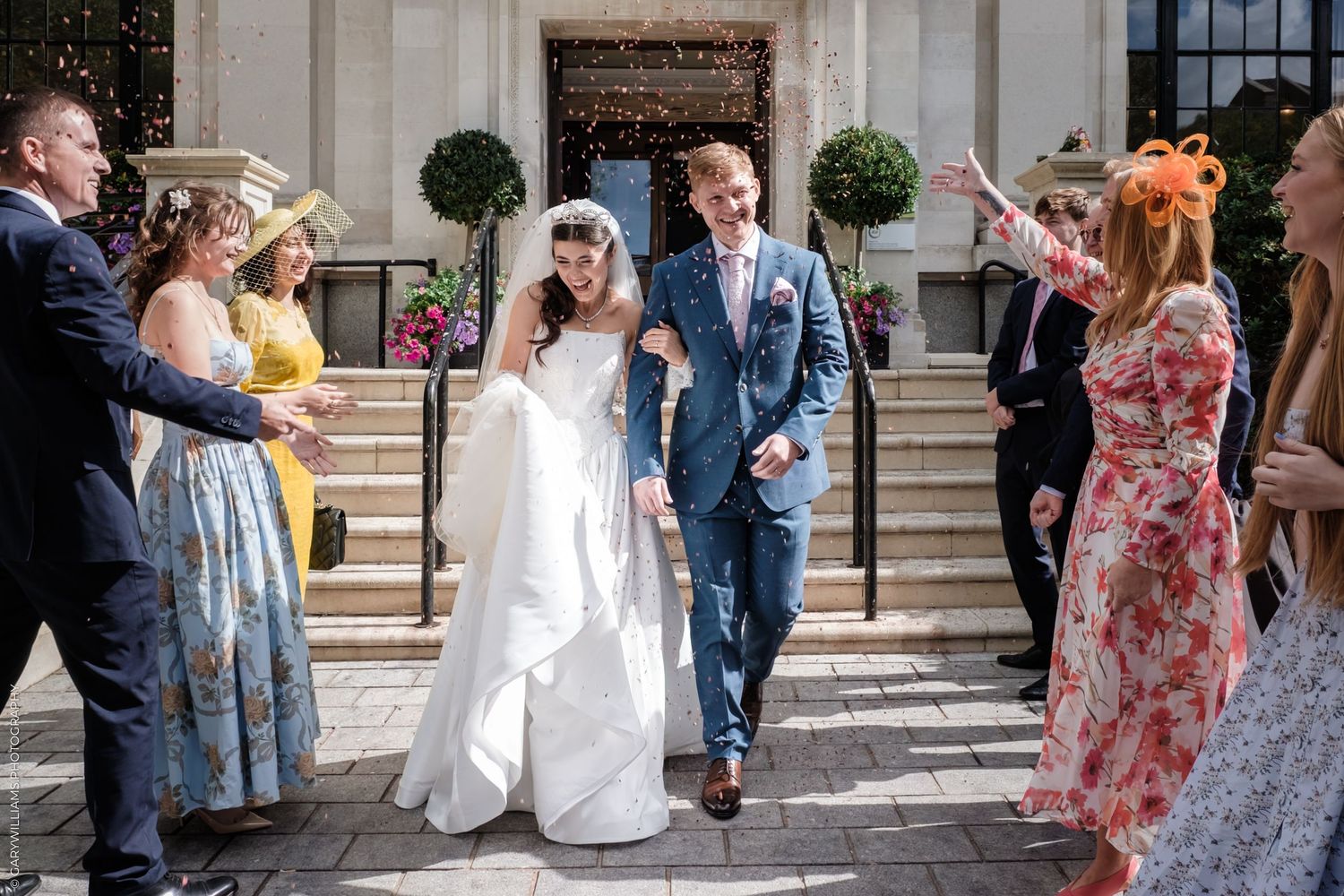 Wedding party celebrating with confetti on stone steps while bride and groom embrace in formal attire.