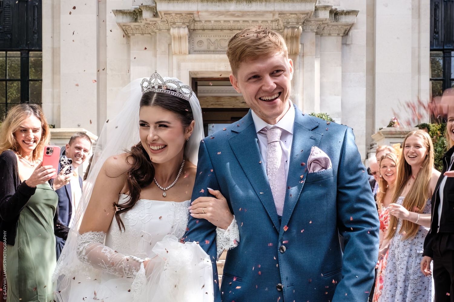 Wedding couple and guests laugh together while standing on grand stone steps of a historic building.
