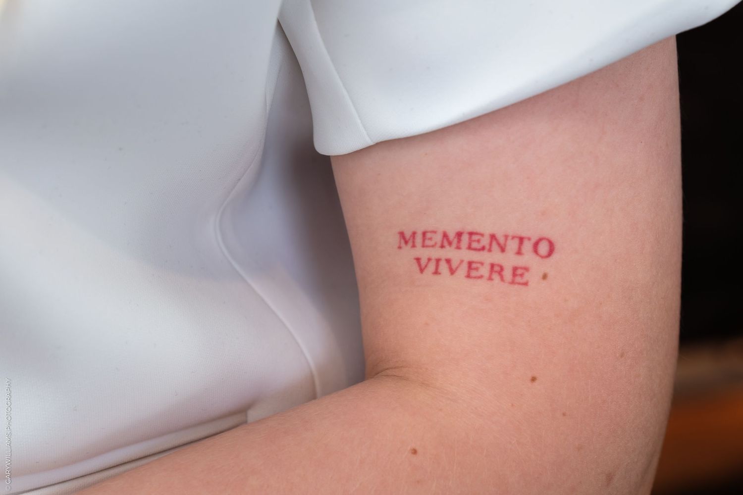 A bare shoulder and arm with a red text tattoo reading 'MEMENTO VIVERE' in serif font.