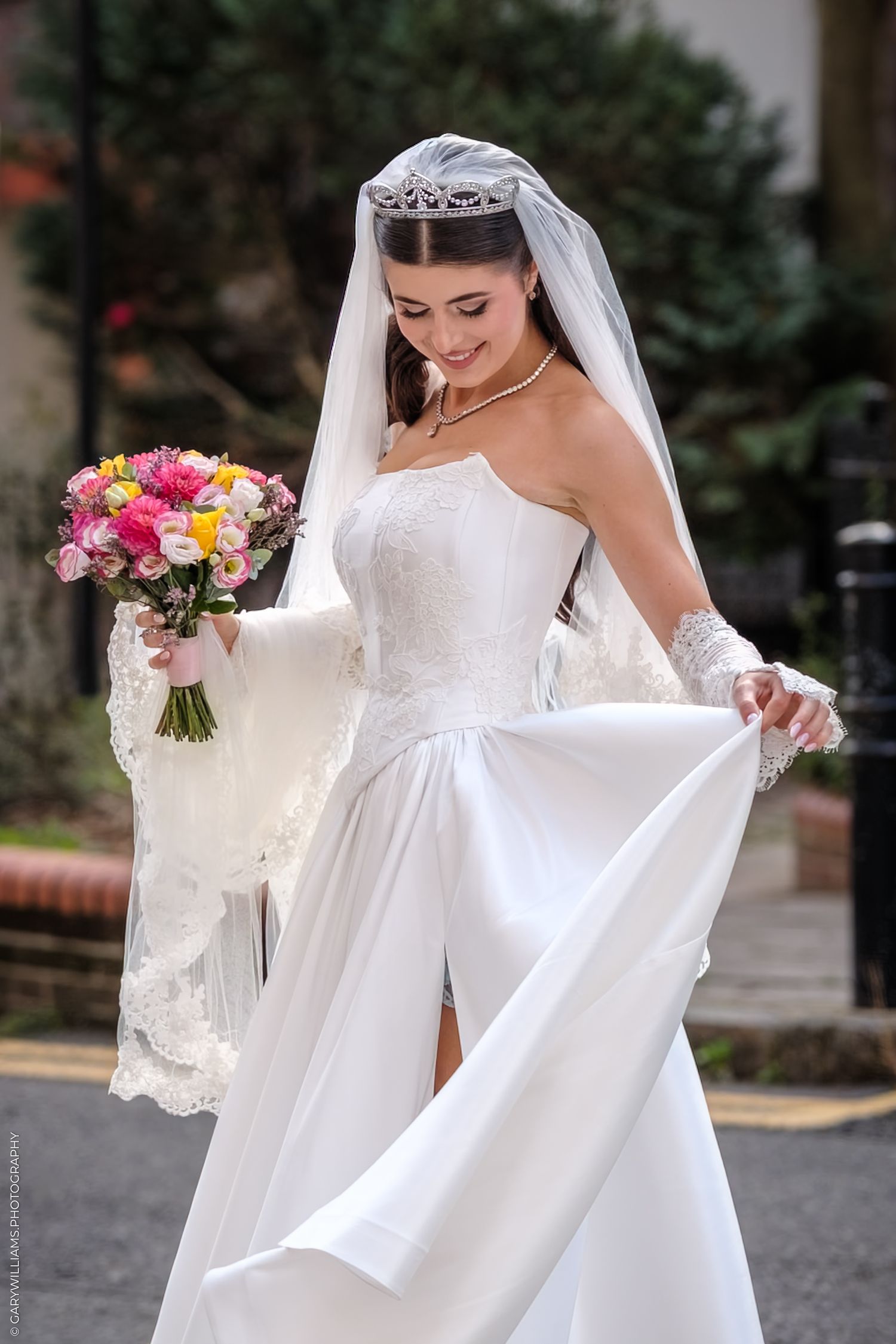 Bride in elegant white wedding gown with flowing veil holds colorful bouquet during outdoor bridal photo session.