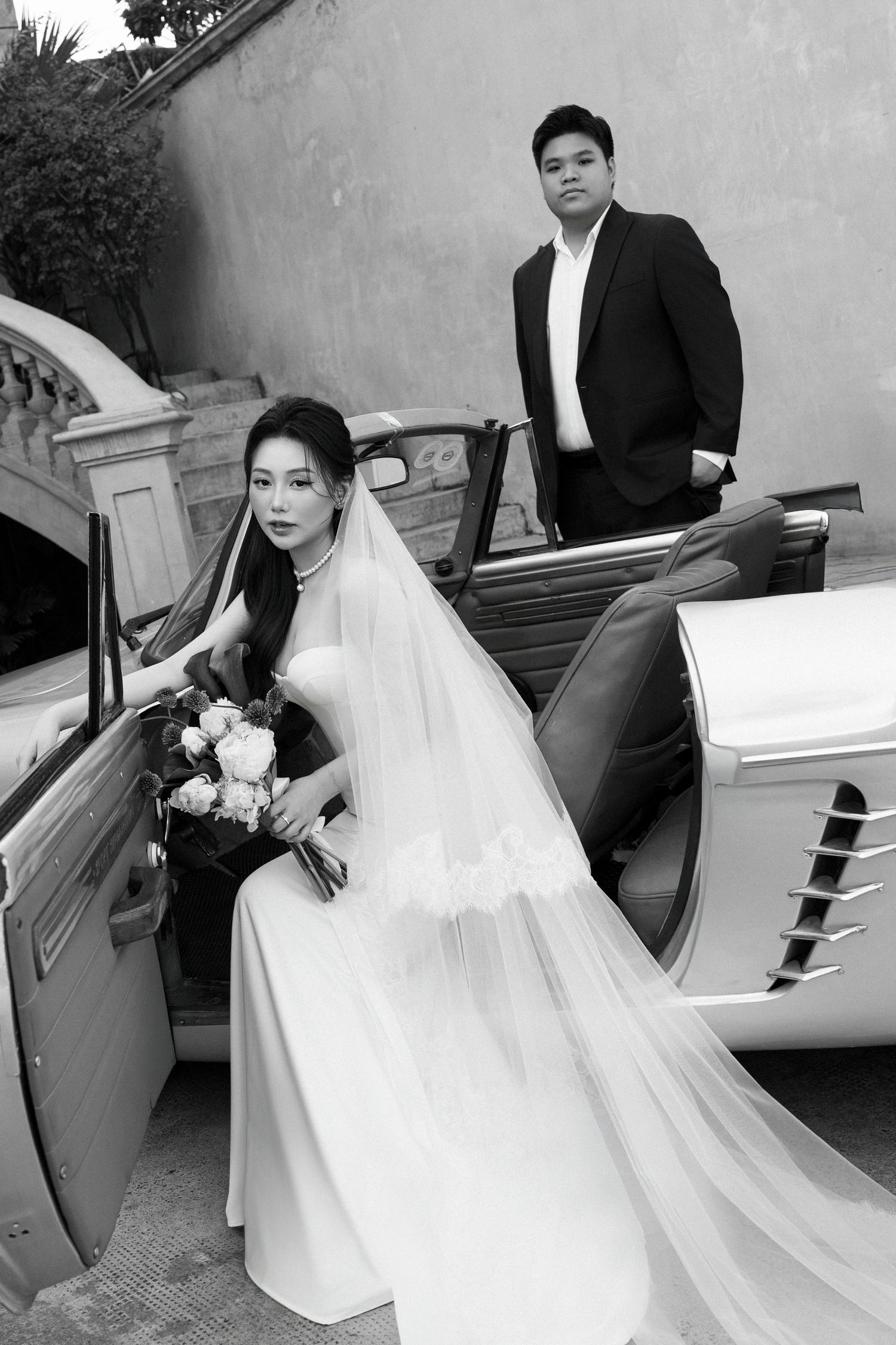 Black and white wedding photo of bride in veil exiting vintage car while groom stands nearby.