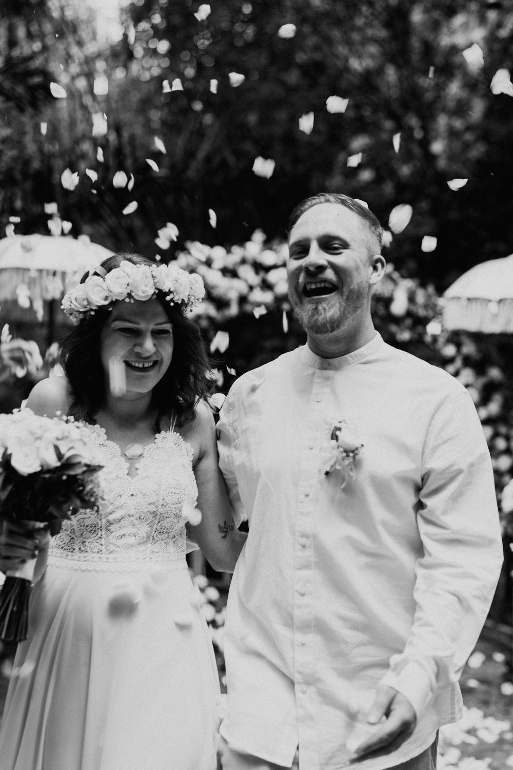 Black and white portrait of a wedding couple laughing together as flower petals fall around them.