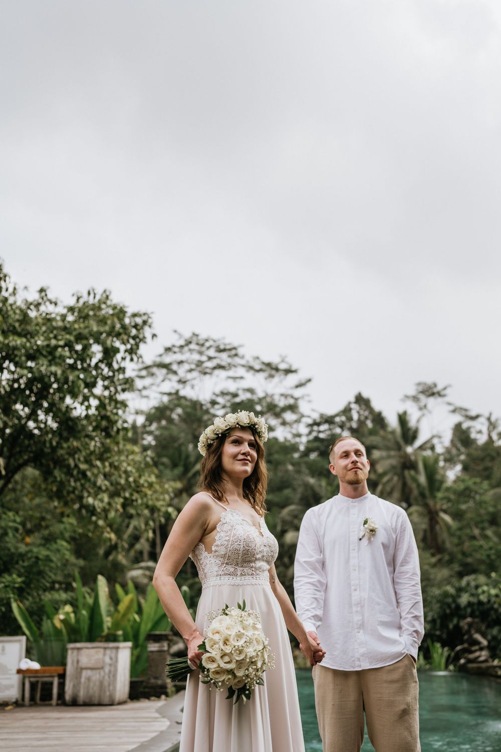 A couple in wedding attire poses together outdoors near a tropical waterfront setting.