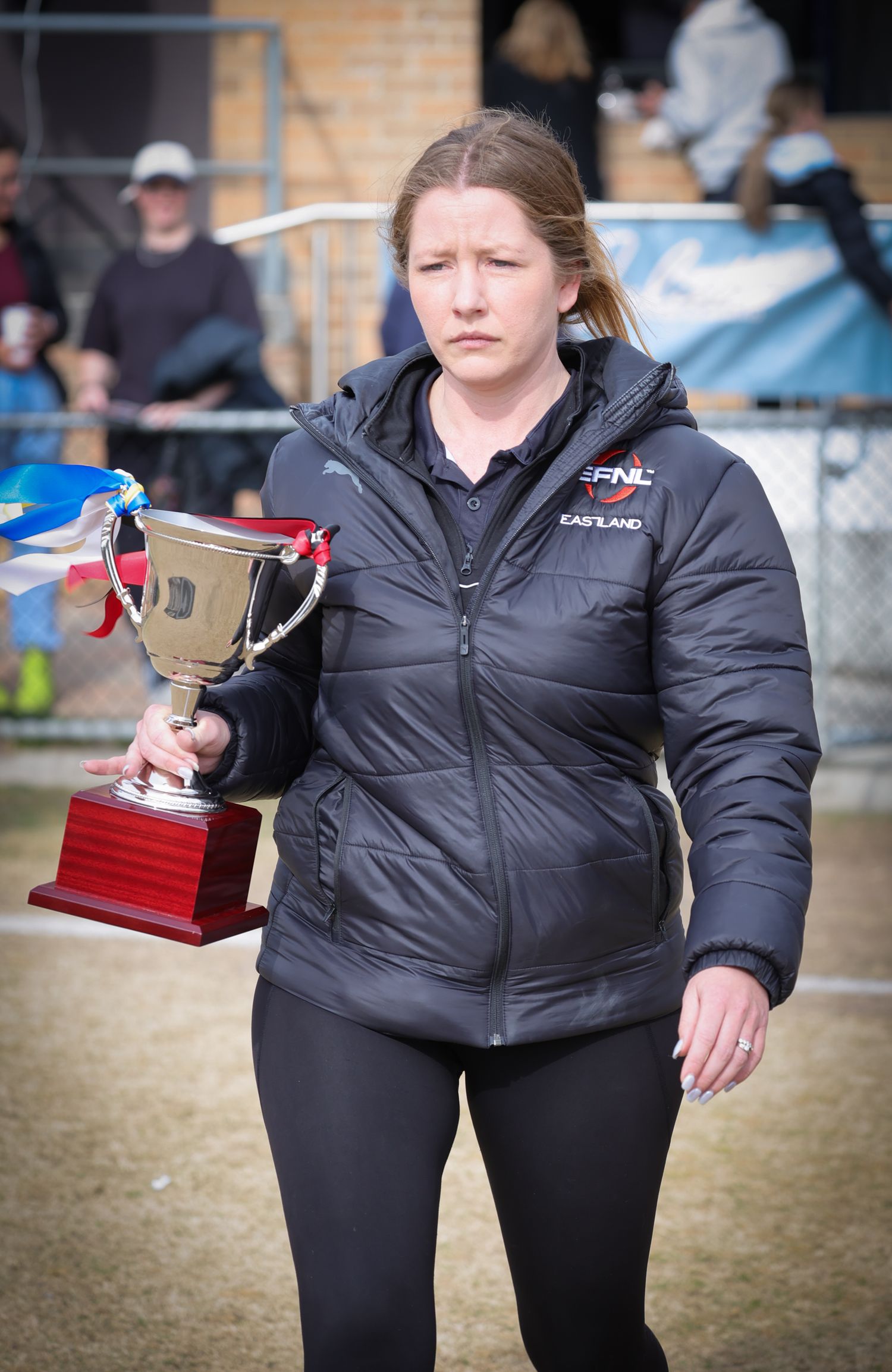 Athlete in black winter jacket carries trophy at outdoor sporting event while walking across field in athletic wear.