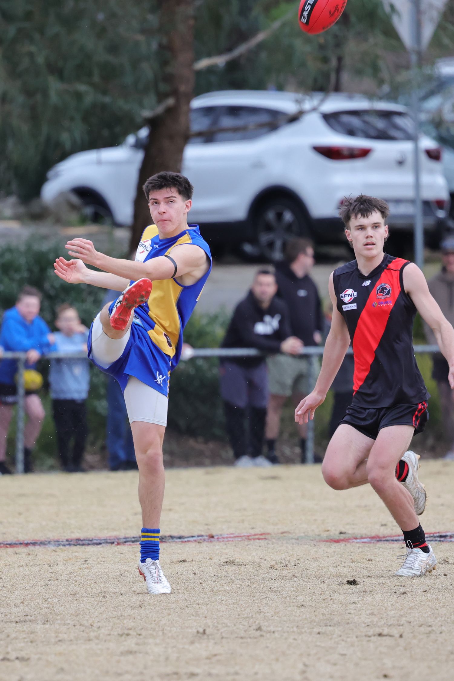 Australian Rules Football player kicks a ball during a match on a grass field with spectators watching.