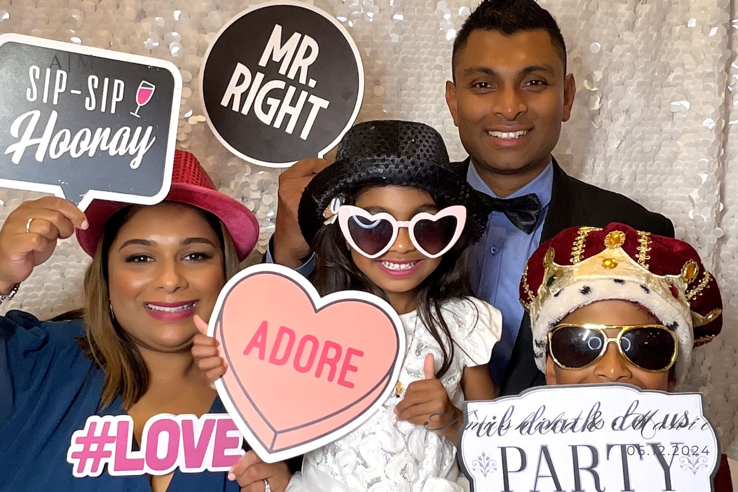 People pose with fun props and signs at a photo booth party wall with glittery backdrop and decorative elements.