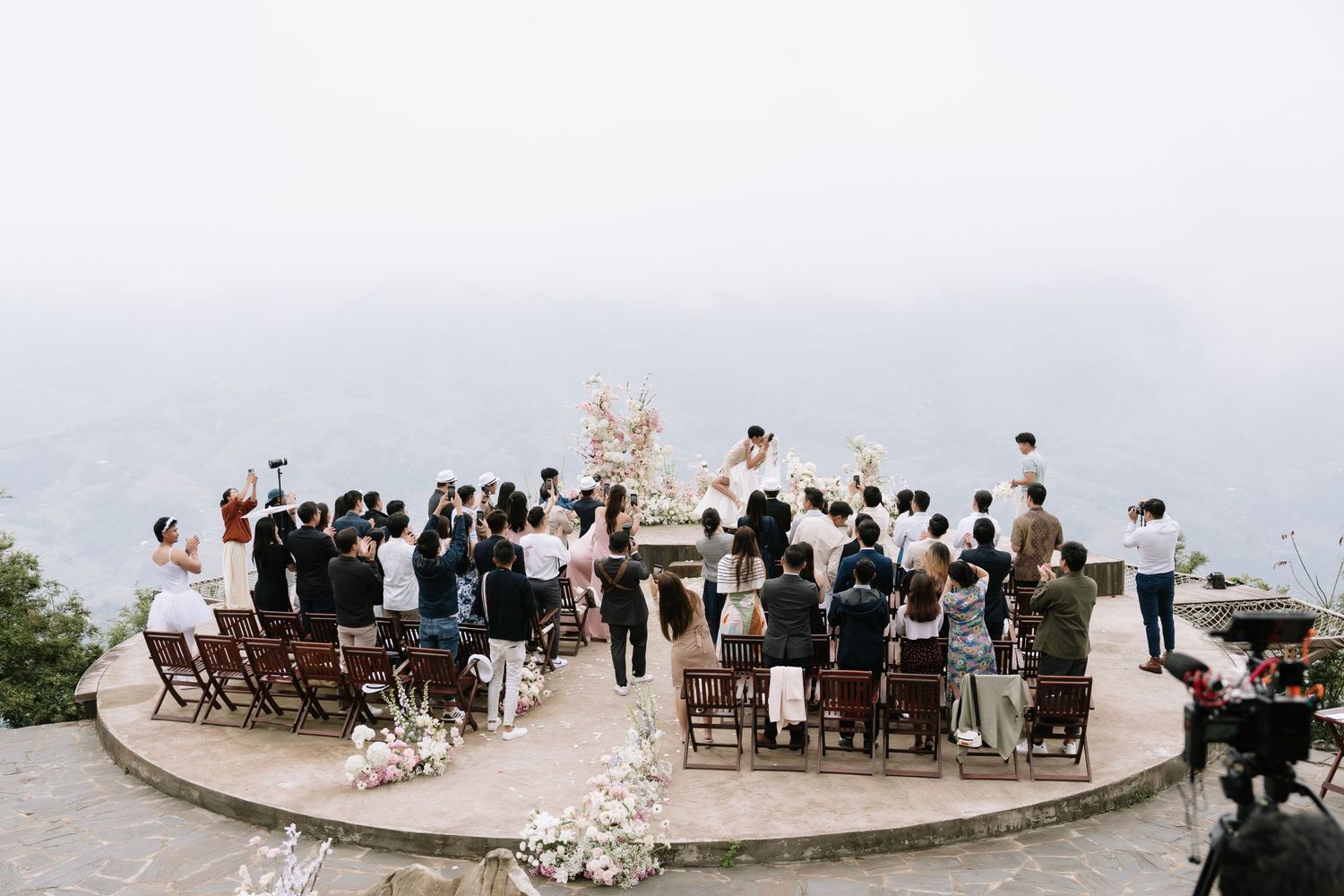Outdoor wedding ceremony on a scenic cliffside terrace with guests seated among floral arrangements and misty mountain views.