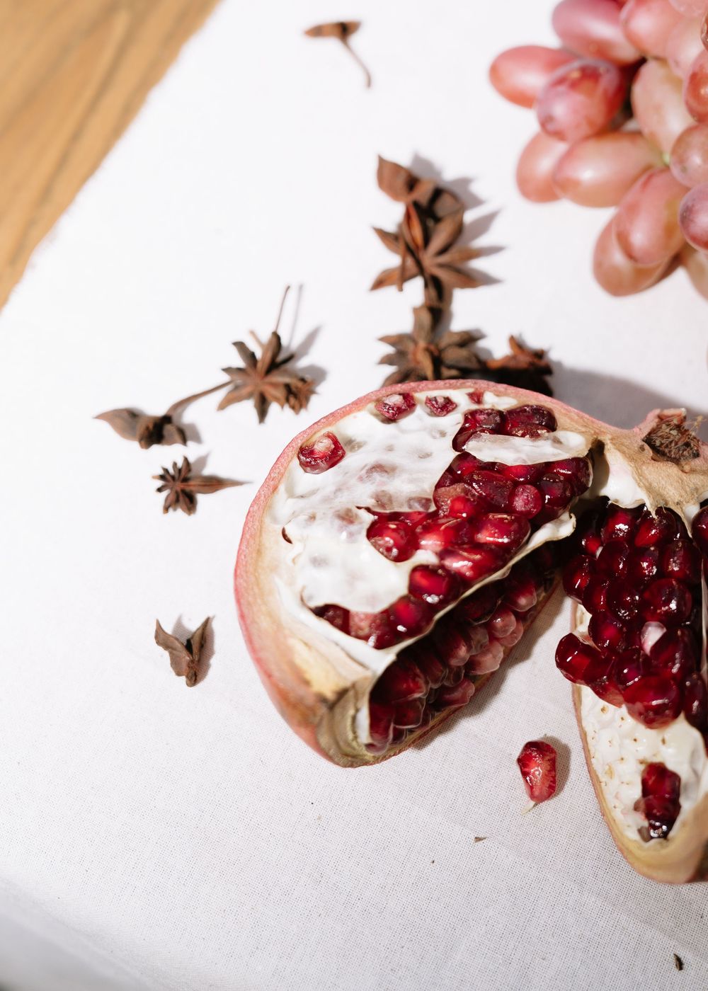 Close-up of a broken pomegranate with red seeds, star anise spices, and grapes on a white surface.