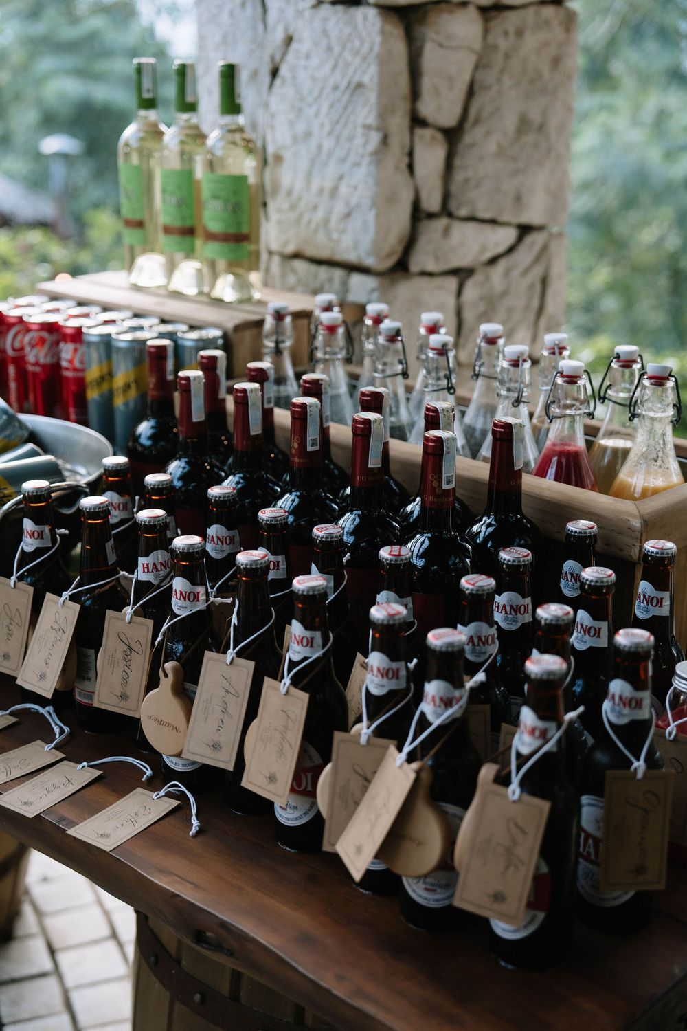 Assorted beer and beverage bottles with gift tags arranged on wooden tables at an outdoor wedding reception.