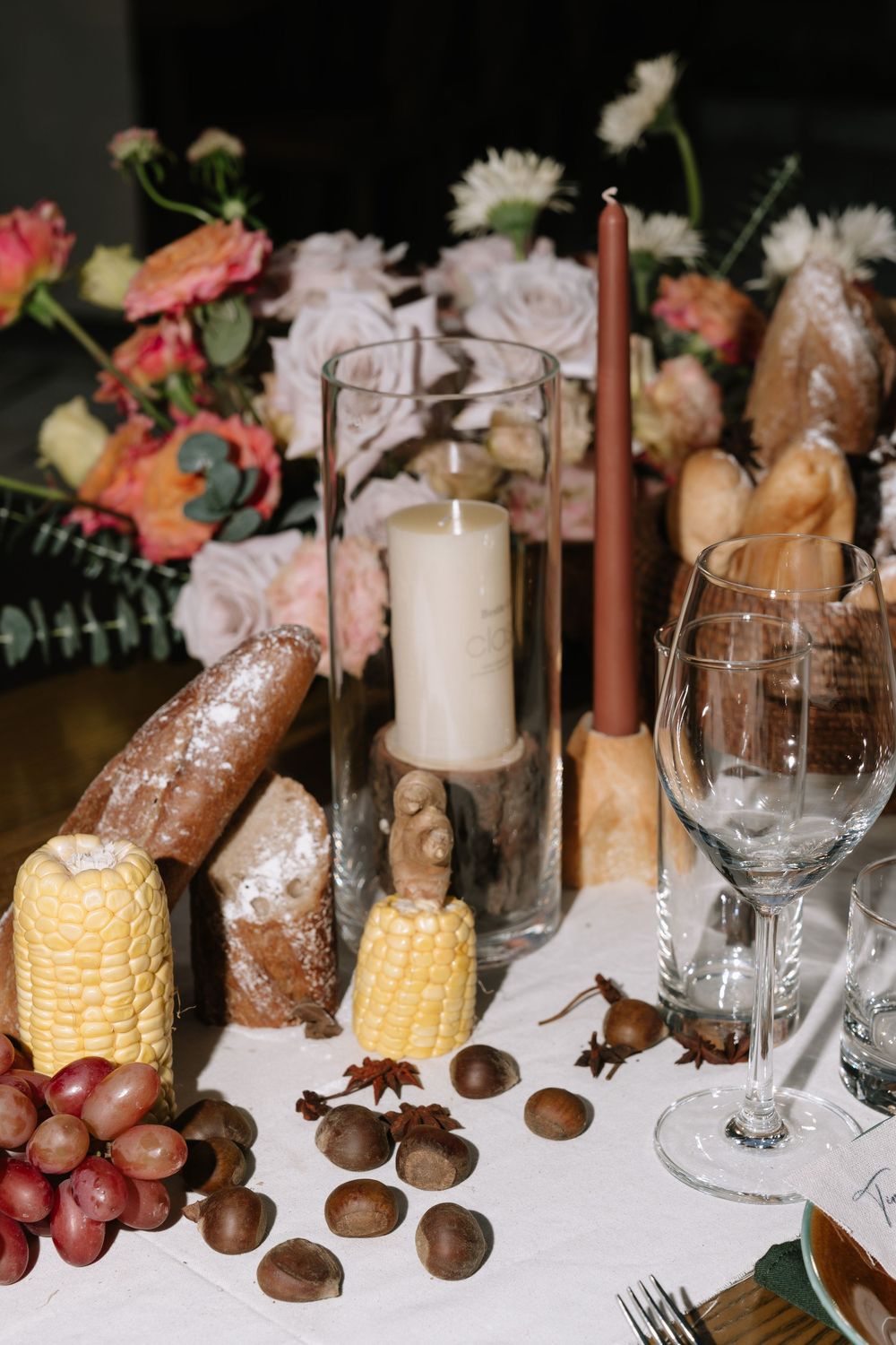 Rustic wedding table centerpiece with candles, bread, corn, chestnuts, grapes, and pink floral arrangement.