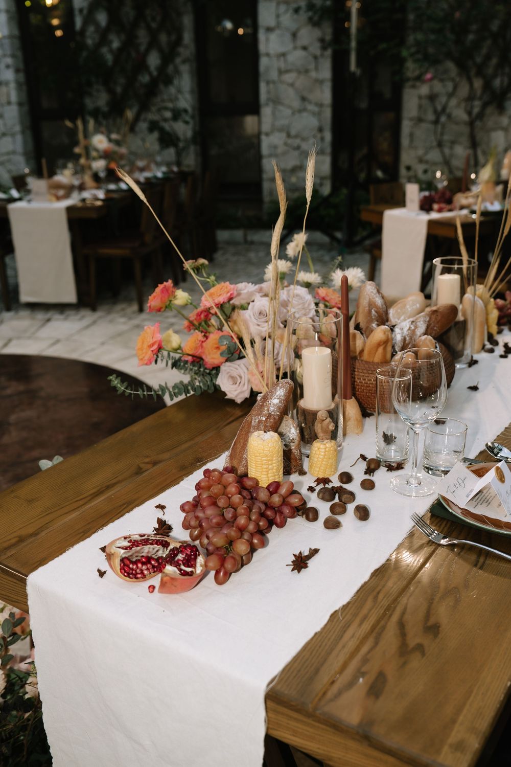 Rustic wooden wedding table with white runner, grapes, pomegranate, corn, chestnuts, candles, and floral centerpiece.