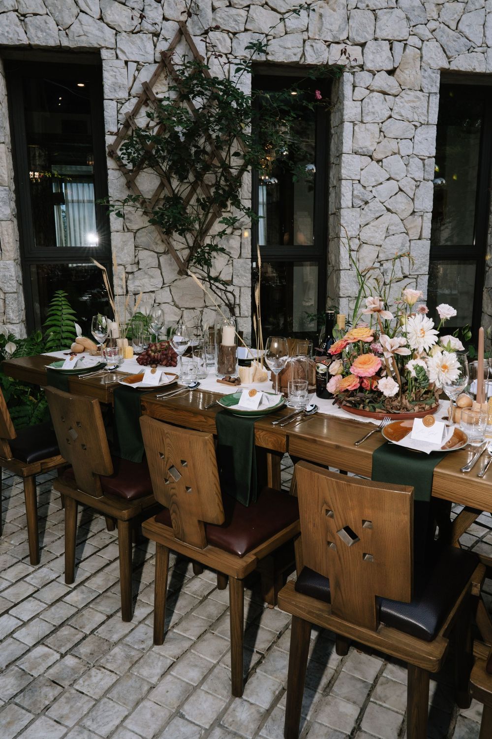 Elegant rustic wedding reception table with wooden chairs, green napkins, floral centerpiece, against stone wall backdrop.