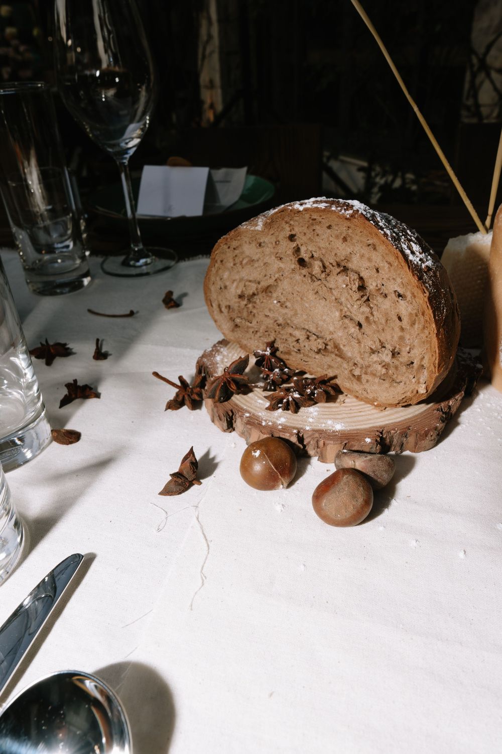 Rustic sourdough bread on wooden board with star anise and hazelnuts on white linen tablecloth with glassware.
