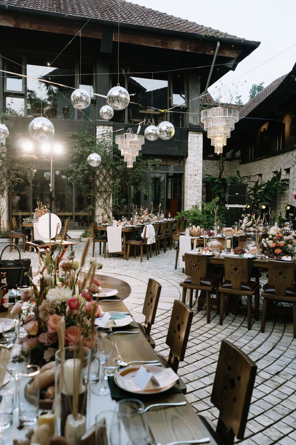 Outdoor wedding reception venue with disco ball chandeliers, floral centerpieces, and wooden chairs at dusk.