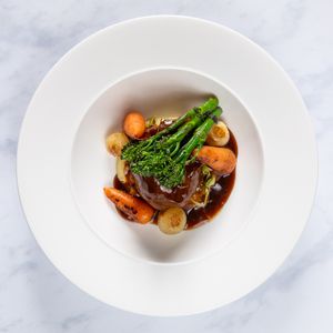 Braised beef with carrots, pearl onions and broccolini served in a white bowl with rich brown sauce on marble surface.