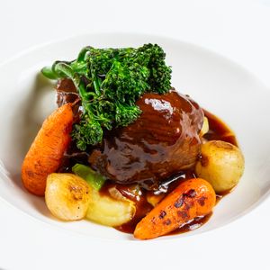 Braised beef short rib with glazed carrots and potatoes in rich brown sauce garnished with fresh parsley.