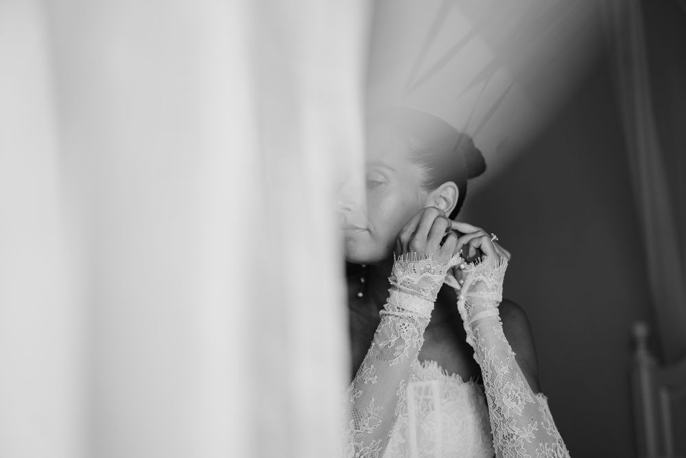 Black and white photo of a bride in lace dress putting on earrings on her wedding day.