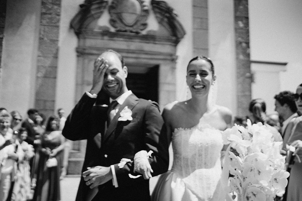 Black and white wedding photo of smiling bride holding bouquet and groom covering face with hand outside church.