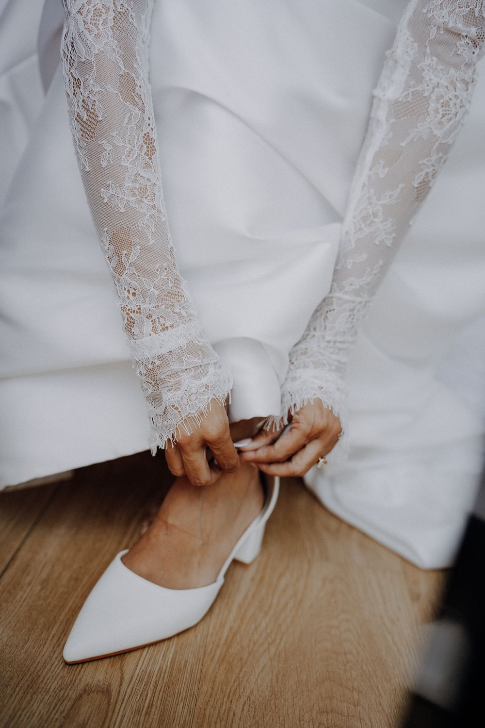 Bride in lace wedding dress adjusting white pointed-toe heels on wooden floor.
