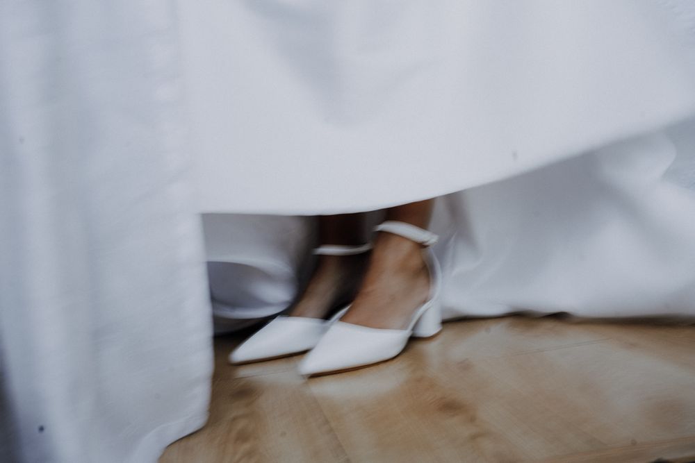 Close-up of a bride wearing elegant white pointed-toe heels with ankle straps beneath a flowing white wedding gown.