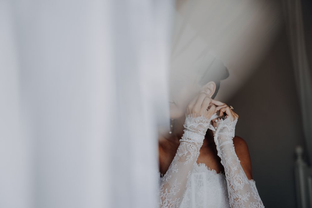 Bride in lace wedding dress and gloves adjusting her veil near a white curtain on her wedding day.