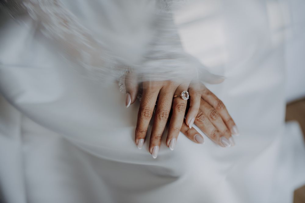 Close-up of bride's hands with elegant white manicure and diamond engagement ring over flowing white wedding dress.