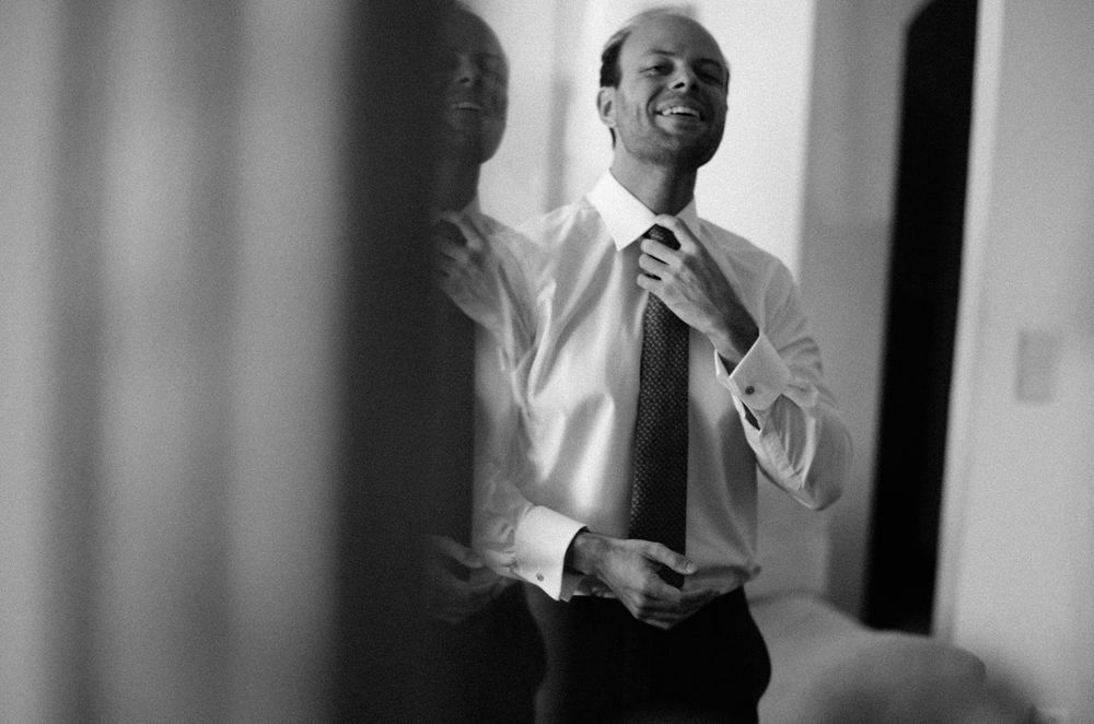 Black and white photo of a smiling man adjusting his tie while getting dressed in a white dress shirt.
