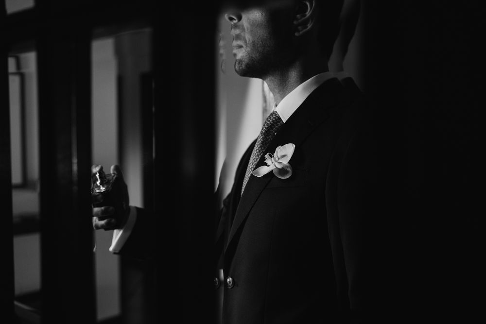 Groom in black suit with boutonniere holding a drink in dramatic black and white wedding portrait.