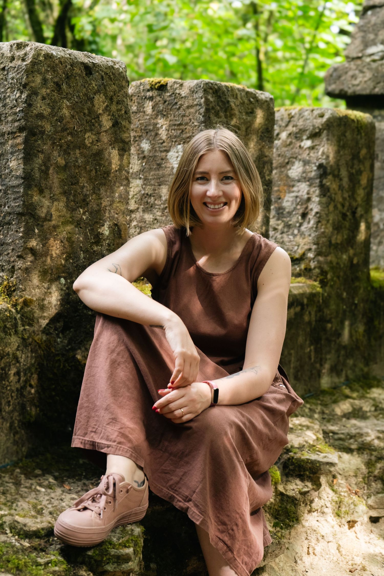 A rustic stone wall setting with female author sat, smiling wearing a brown dress and casual shoes.