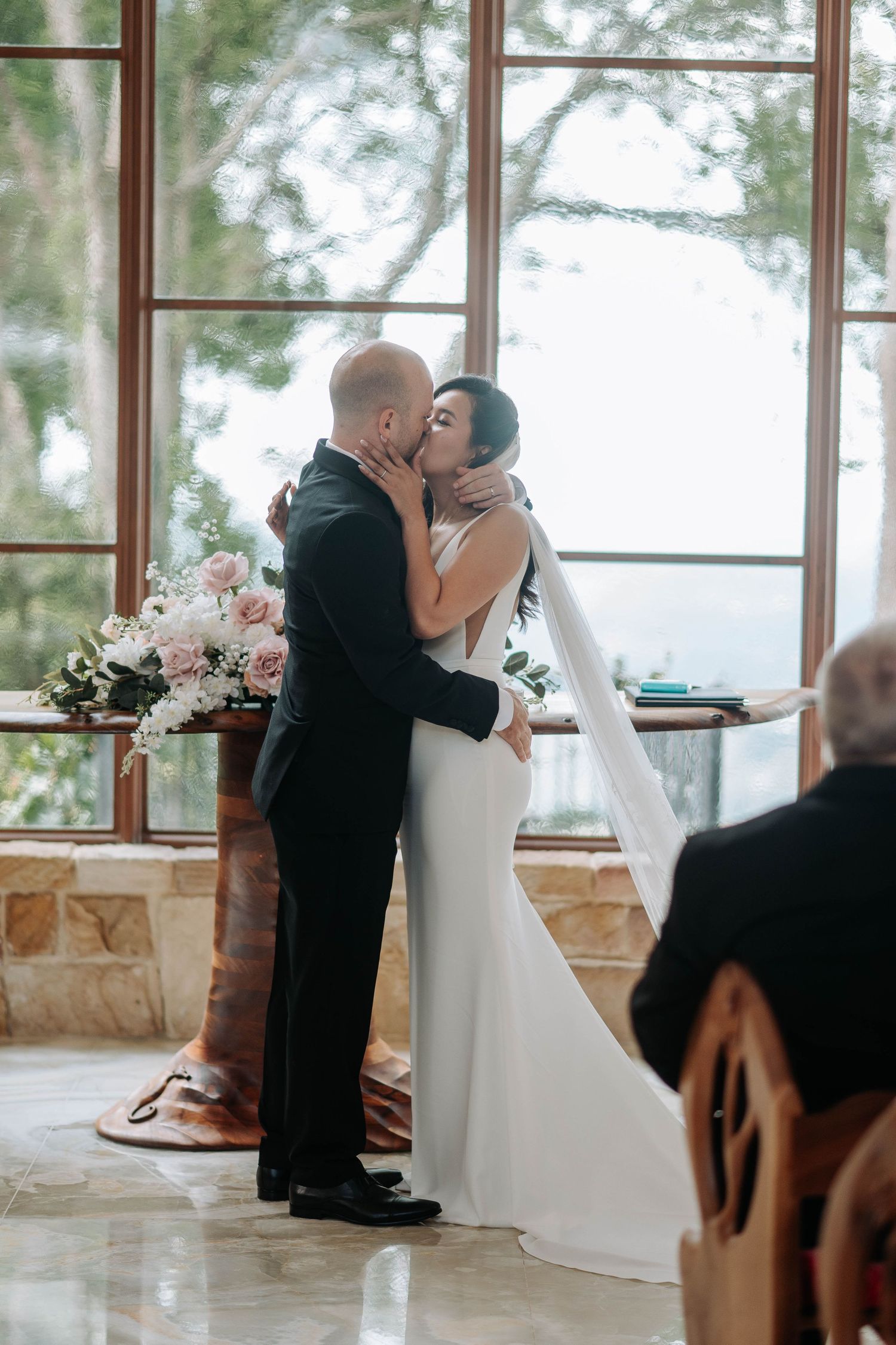 A couple shares their first kiss as newlyweds during an intimate wedding ceremony in a bright sunlit room.