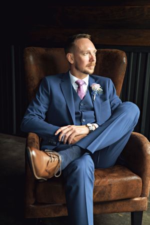 A groom in a navy blue suit relaxes on a brown leather couch in moody lighting.