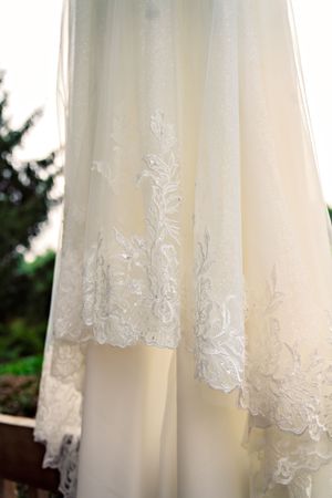 Close-up detail of delicate lace embroidery on the hem of an ivory wedding dress veil against a blurred outdoor background.