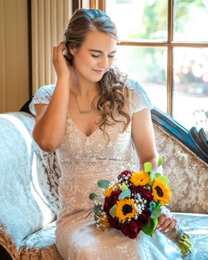 Bride holding vibrant fall wedding bouquet with sunflowers and burgundy flowers against white lace dress in natural light.