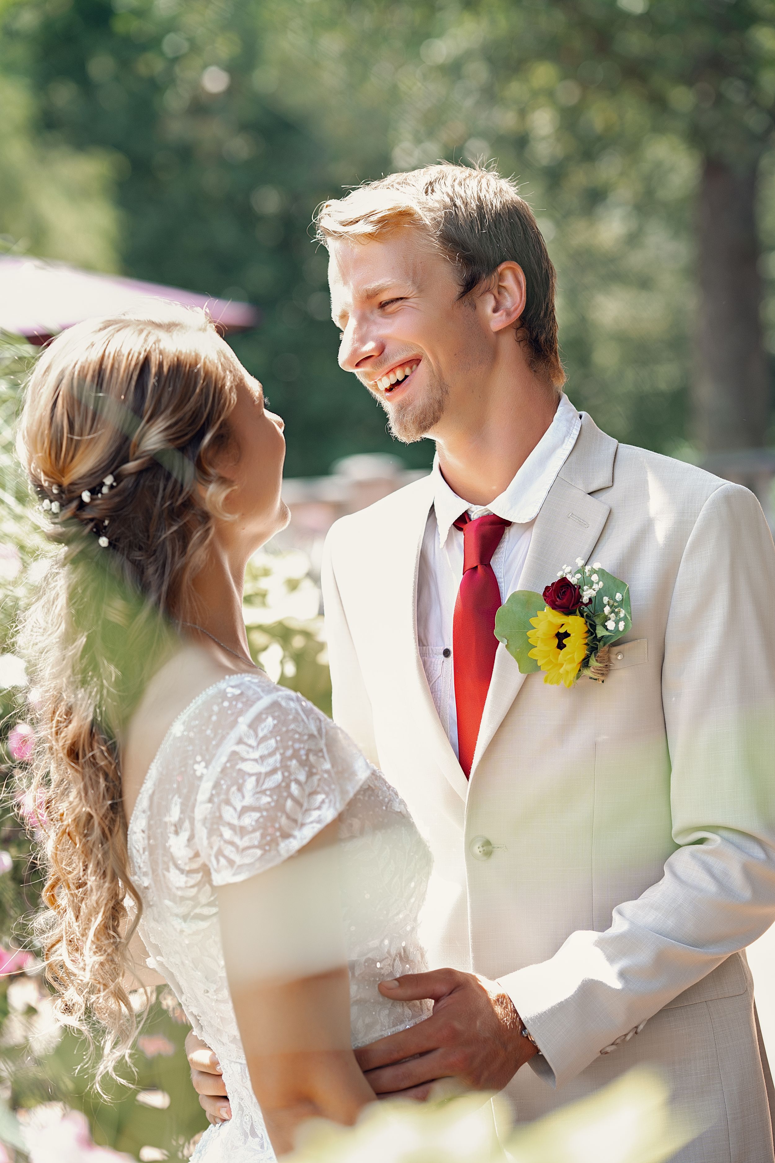 Joyful newlyweds share intimate moments outdoors in natural sunlight wearing elegant white and red formal attire.