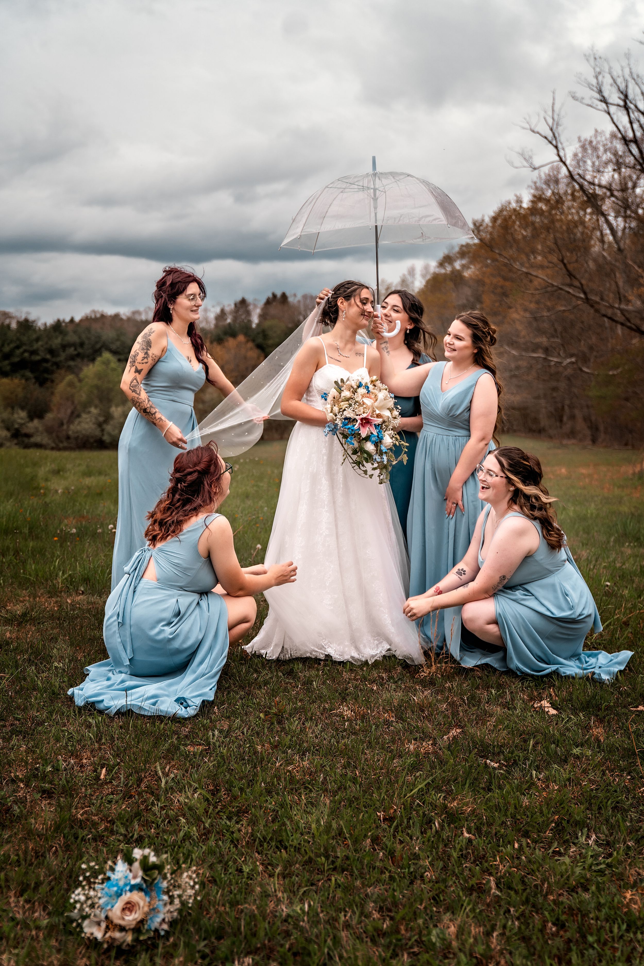 A bride in white stands with bridesmaids in light blue dresses in a field under stormy skies with a clear umbrella.