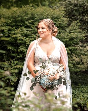 A bride in a white lace wedding dress holds a beautiful bouquet of blush and blue flowers with eucalyptus in an outdoor setting.