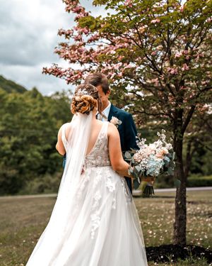 A romantic wedding couple holds hands beneath pink cherry blossoms on a cloudy spring day.