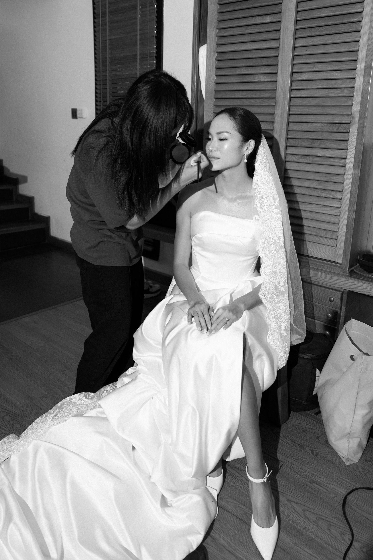 Black and white photo of makeup artist applying makeup to seated bride in white gown.