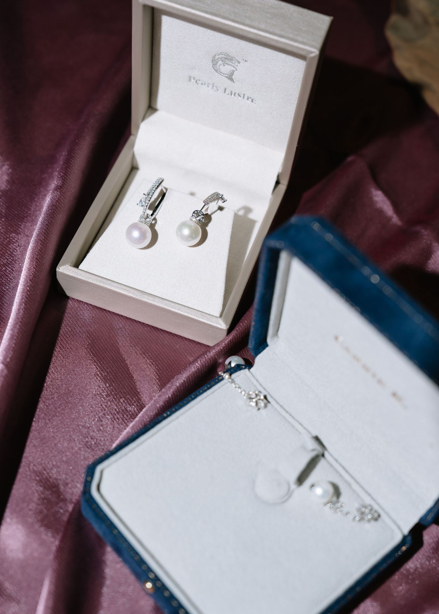 Pearl drop earrings and necklace in luxury jewelry boxes on purple satin fabric for wedding day.