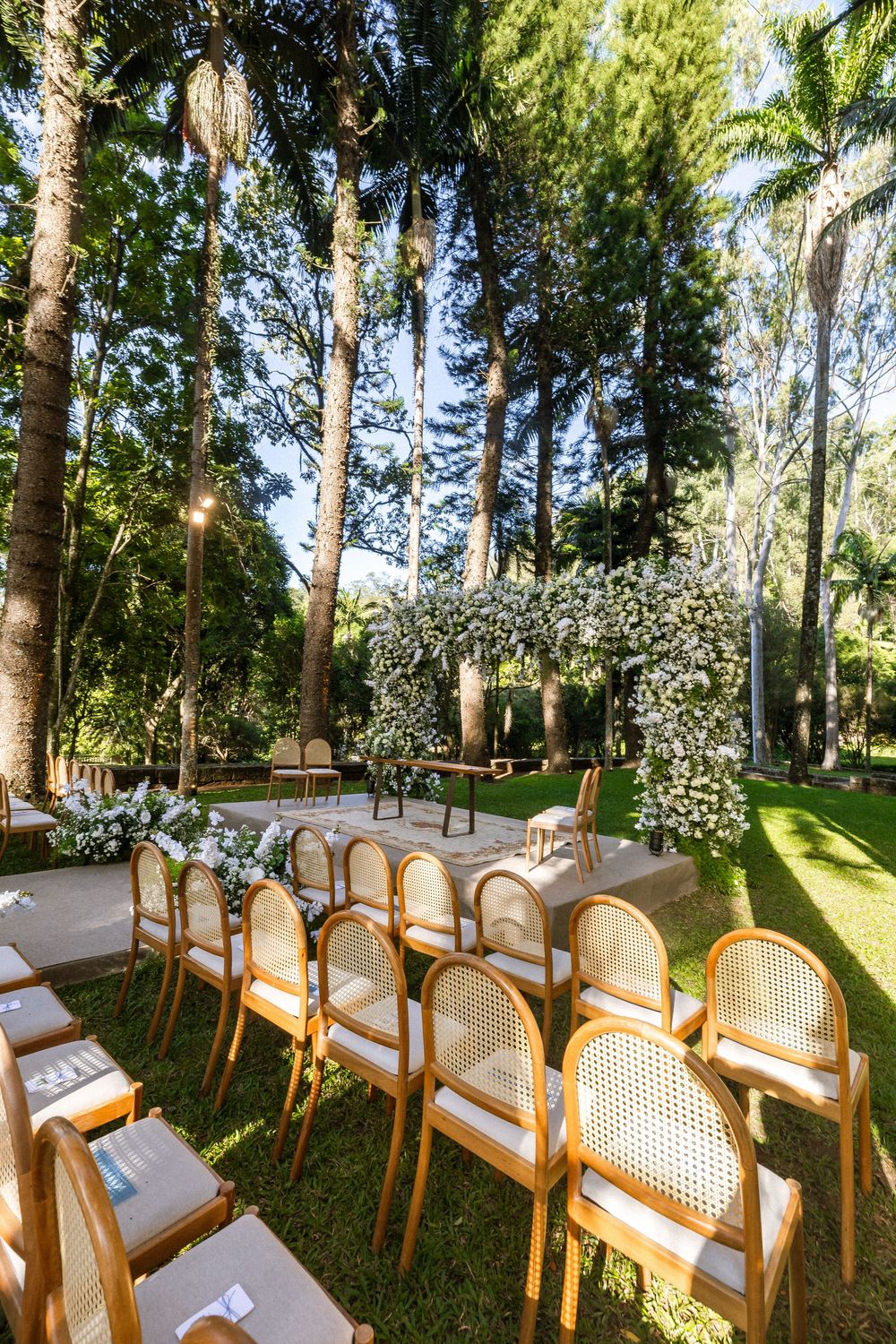 Wooden chairs with cane backs and white cushions arranged in rows for an outdoor wedding ceremony under tall trees.