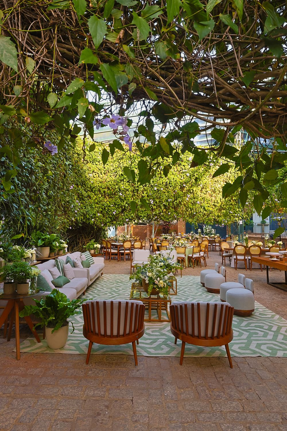 Indoor-outdoor lounge area with wooden chairs and grey cushions set under leafy canopy and geometric pattern rug.