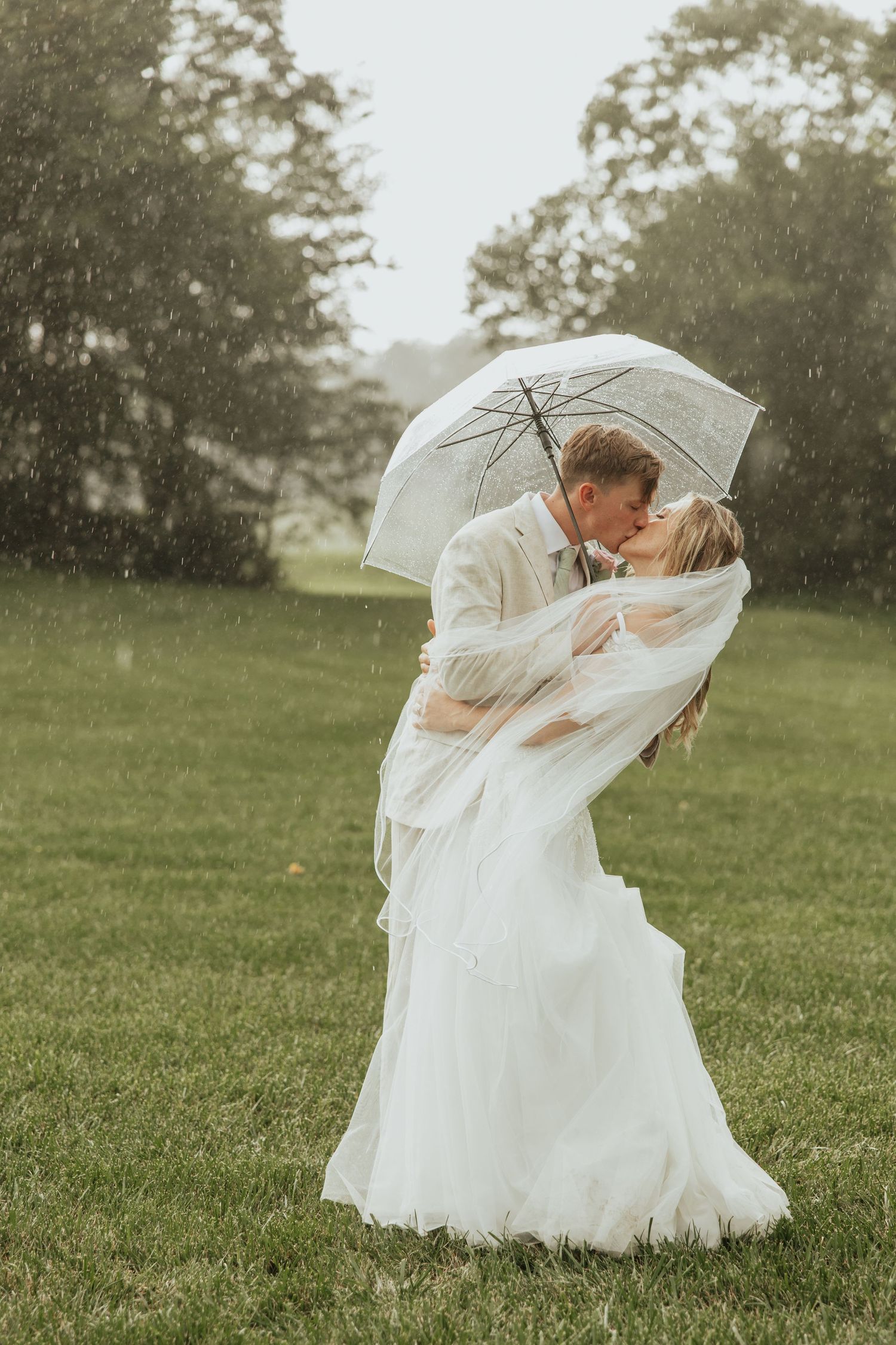 Romantic couple shares intimate kisses under a white umbrella in the rain during outdoor wedding photography session.