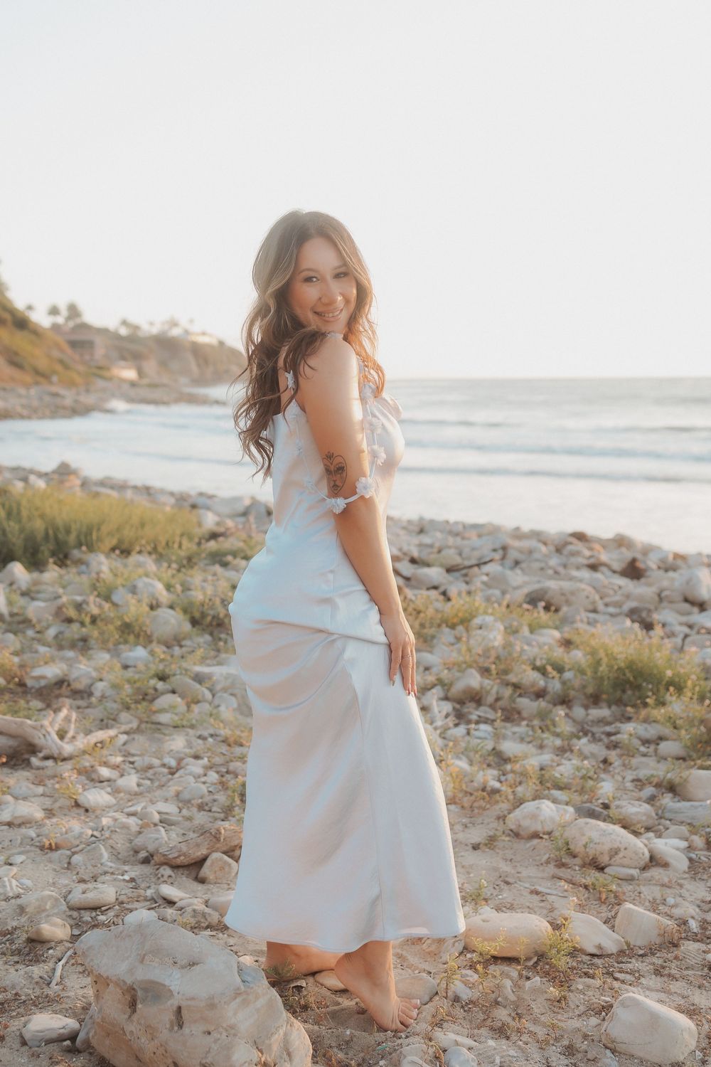 Person in a flowy white dress poses on a rocky beach at sunset with ocean waves in the background.