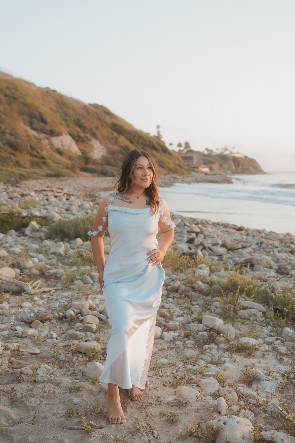 A woman wearing a flowy white dress poses gracefully along a rocky coastal beach during golden hour lighting.