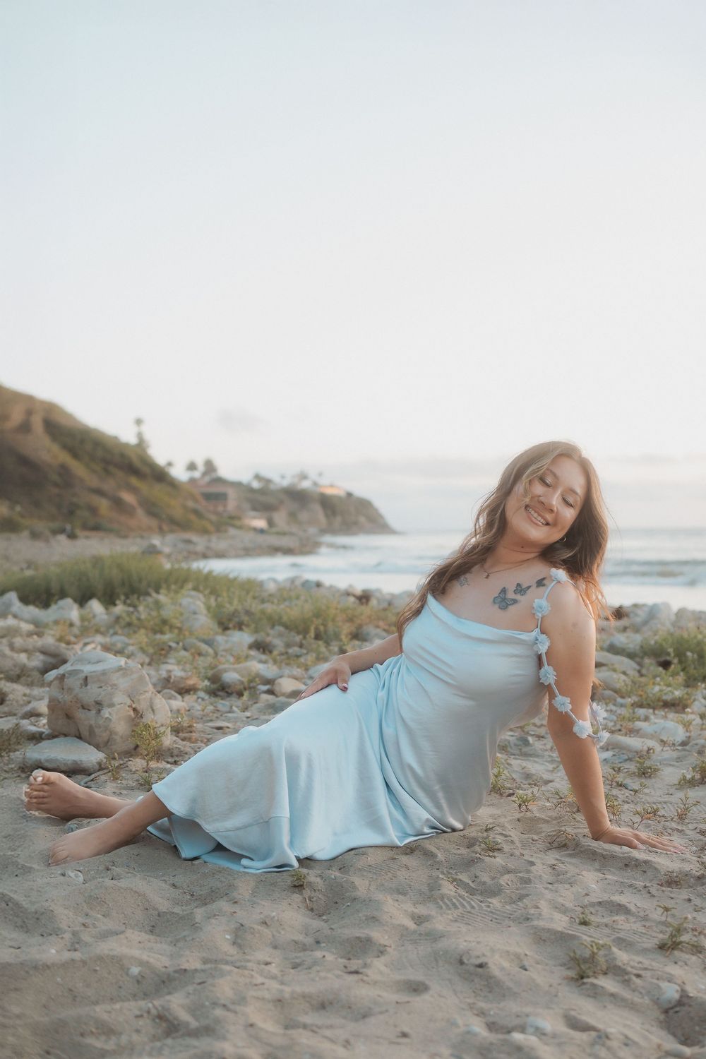 A light blue dress flowing in the breeze as someone poses on a rocky beach at sunset with cliffs in the distance.