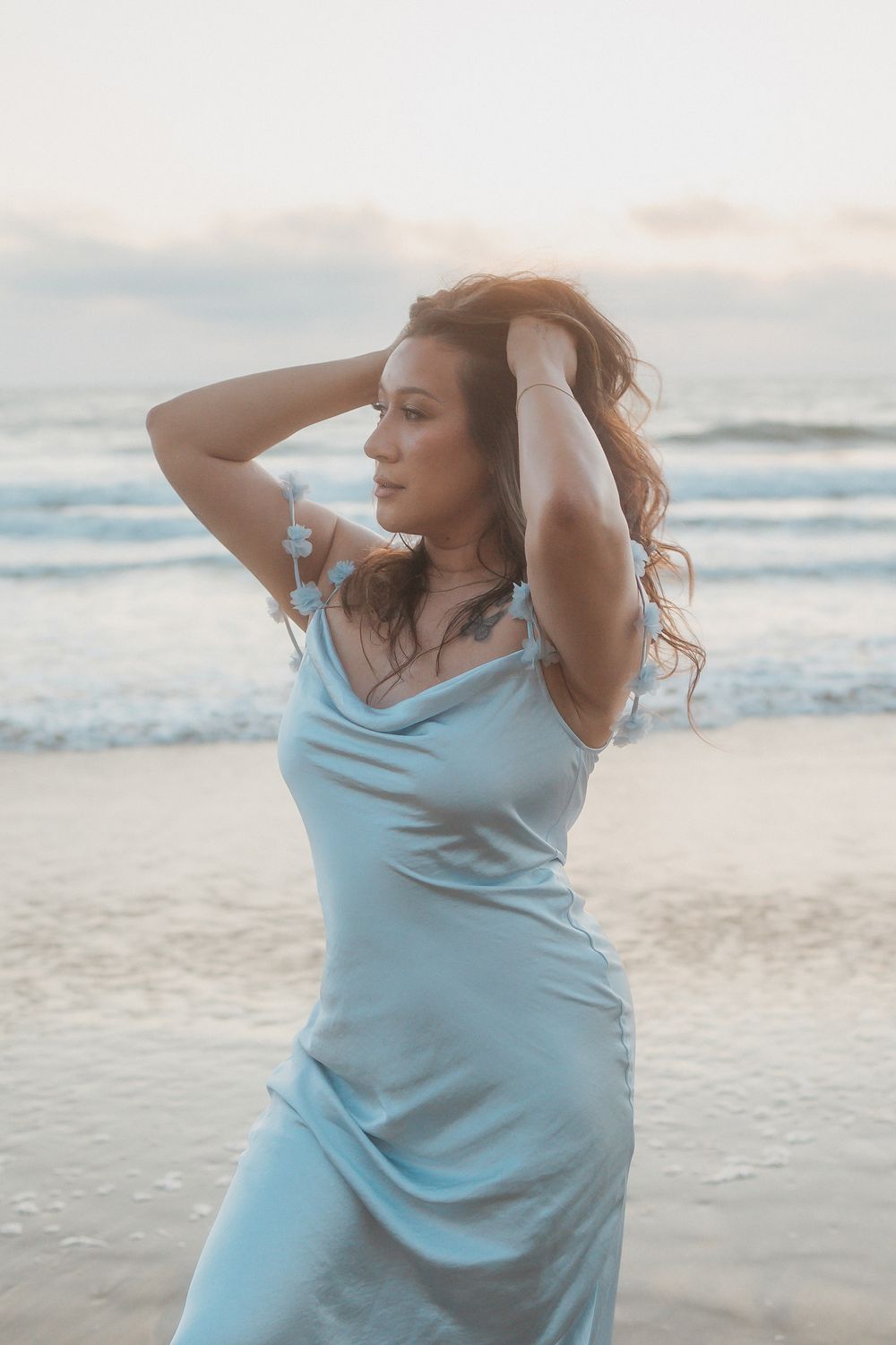 Person in a light blue tank top poses artistically on a serene beach at sunset with ocean waves in the background.